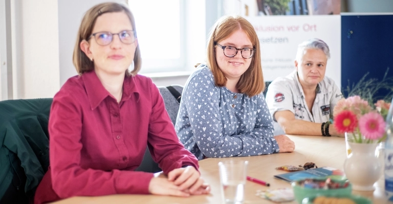 Drei Frauen sitzen an einem Tisch mit Blumen und lächeln.