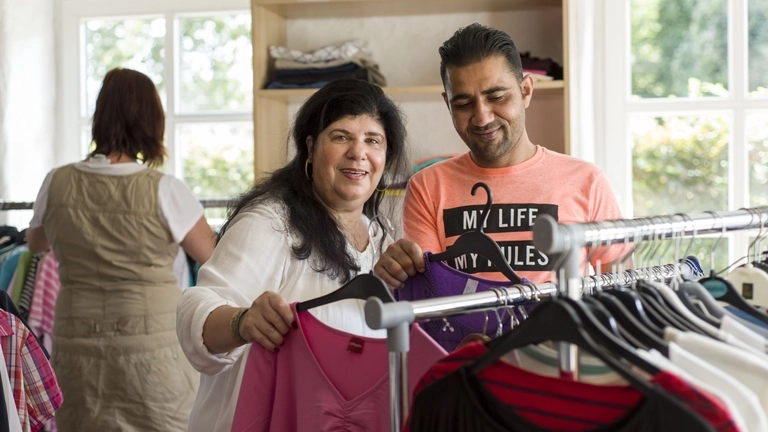 Eine Frau und ein Mann sortieren Kleindung an einem Kleiderständer in einer Kleiderkammer.
