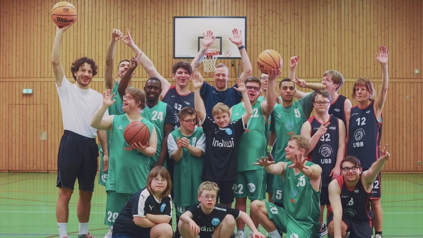 Etwa 20 junge Menschen in grünen und blauen Basketballtrikots  haben sich zu einem Gruppenfoto im Mittelkreis einer Sporthalle aufgestellt. Die Jüngsten sind unter 10, die Ältesten etwa 30 Jahre alt. Alle winken in die Kamera.