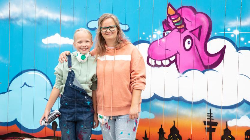 Ein Mädchen und eine Frau stehen Arm in Arm nebeneinander. Sie tragen farbbespritzte Kleidung. Sie stehen vor einer Wand mit einem Einhorn-Graffiti.