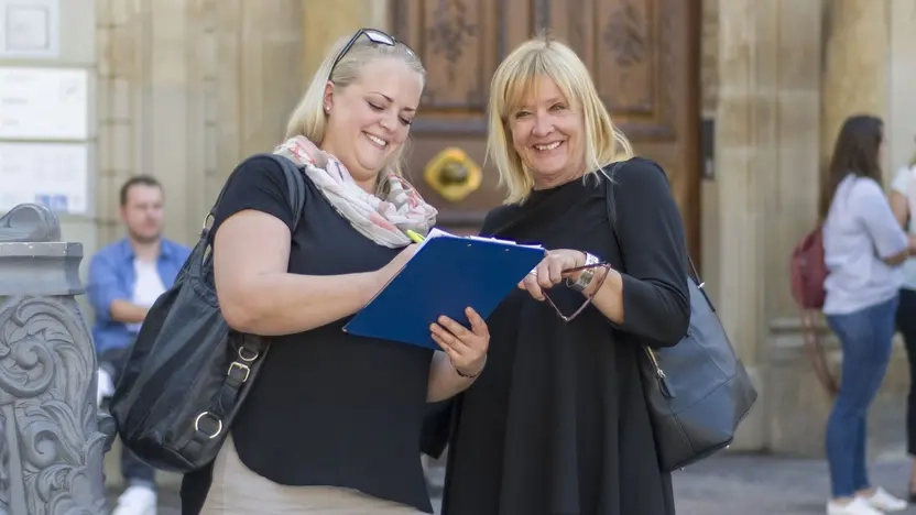 Zwei Frauen stehen vor einem Gebäude und füllen etwas auf einem Klemmbrett aus.