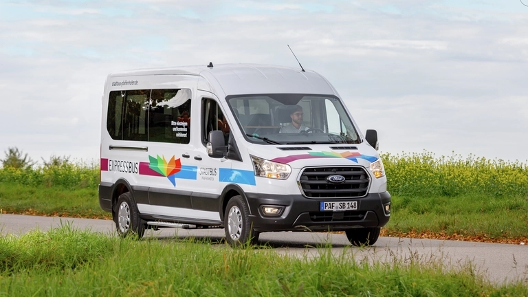 Ein kleiner Personen-Bus fährt über eine Landstraße, umgeben von Bäumen und Wiesen.