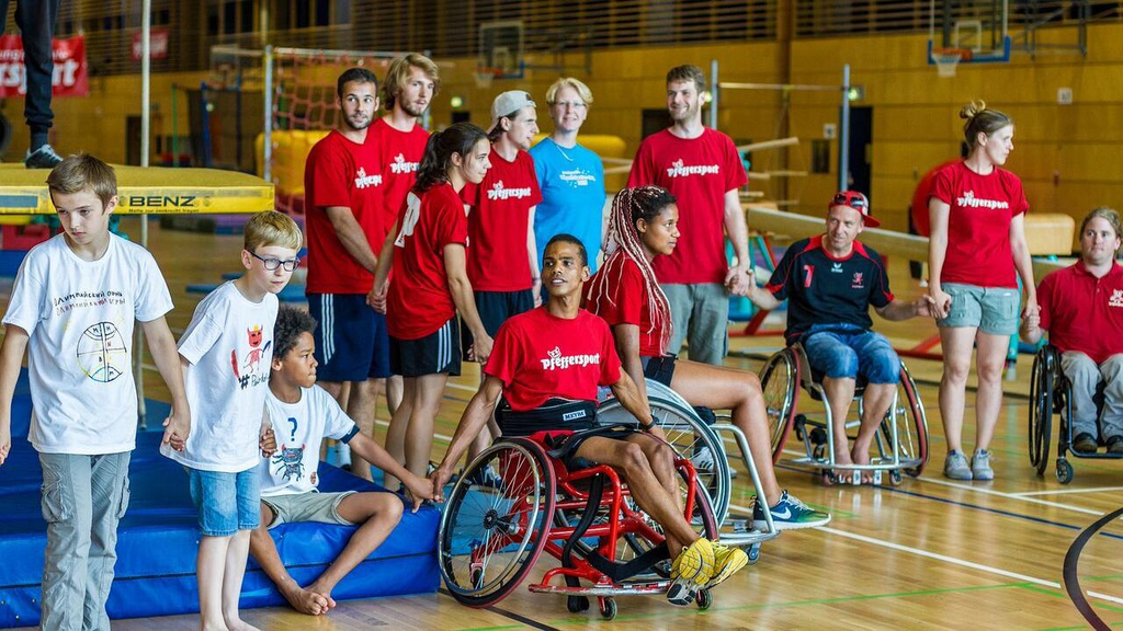 Eine Gruppe Menschen hält sich in einer Reihe an den Händen. Manche sitzen im Rollstuhl. Viele tragen ein rotes T-Shirt mit der Aufschrift "Pfeffersport".
