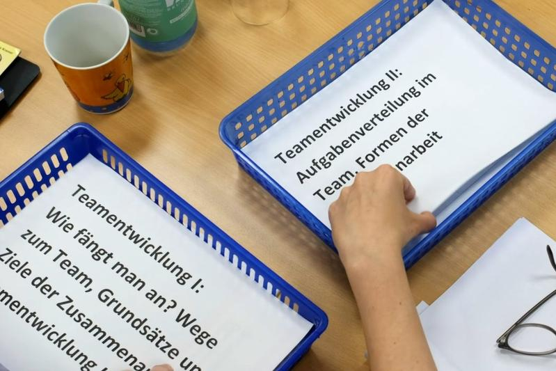 Auf einem Tisch stehen zwei blaue Plastikkörbe. In ihnen liegen Zettel zur Teamentwicklung. Eine Hand greift nach einem der Zettel.