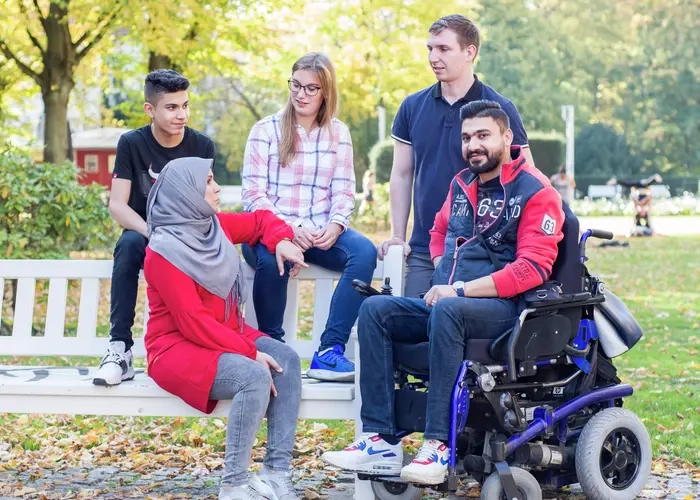 Eine Gruppe von Menschen sitzen im Park, eine Person sitzt im Rollstuhl, eine trägt ein Kopftuch.
