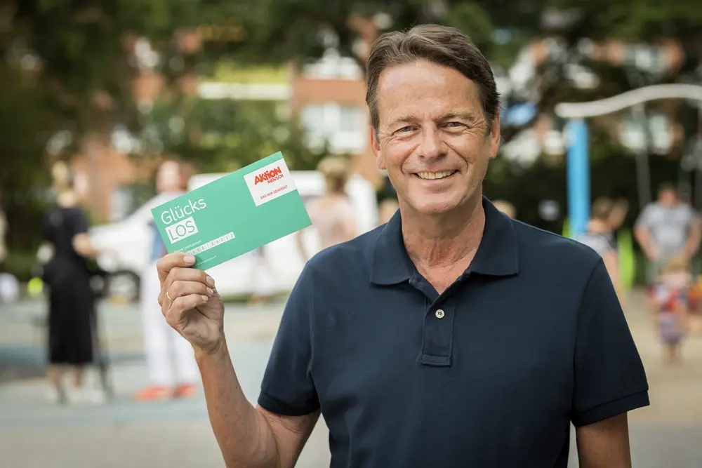 Rudi Cerne trägt ein dunkelblaues Polohemd. Er lacht in die Kamera und hält mit der rechten Hand das grüne Glücks-Los der Aktion Mensch hoch.