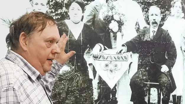 Ein mittelalter Mann in kariertem Hemd steht vor einem s/w-Foto einer Familie und spricht darüber