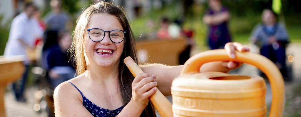 Eine junge Frau mit Down-Syndrom hält eine Gießkanne im Spaß wie ein Megaphon an ihren Mund. Sie lacht fröhlich in die Kamera.