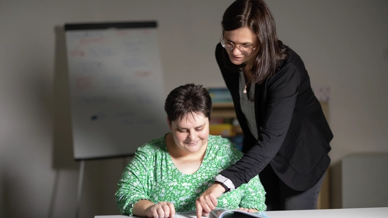 Zwei Frauen in einem Seminarraum: Die eine sitzt an einem Tisch, die andere steht hinter ihr und zeigt ihr etwas in einem Heft.
