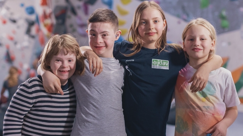 Vier Kinder mit und ohne Behinderung stehen nebeneinander vor einer Boulderwand