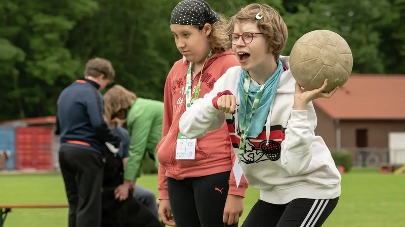 Zwei Mädchen stehen auf einem Sportplatz. Eine von ihnen ist kurz davor, einen Ball zu werfen. Dabei ruft sie etwas.