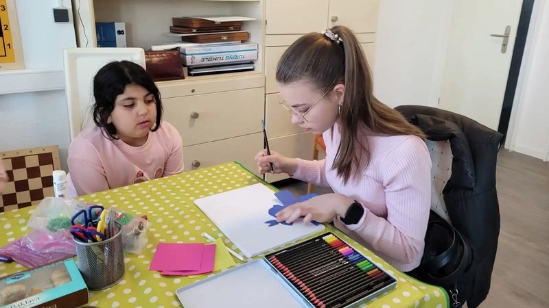 Zwei Mädchen sitzen an einem Tisch. Die Ältere hat einen Zeichenblock vor sich und einen Schere in der Hand. Die Jüngere schaut ihr zu.