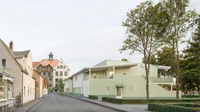 An einer ruhigen Straße ist das Modell eines langgestreckten flachen Neubaus mit geräumigen Balkons und einer hellgrünen Fassade zu sehen. Neben und hinter dem Haus stehen hohe Bäume.