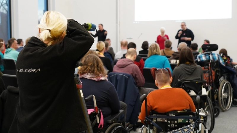 In einem Saal sitzen viele Zuhörer, die von hinten zu sehen sind, darunter auch einige Rollstuhlnutzer*innen. Vorne stehen ein Mann und eine Frau mit Mikrophonen vor den Zuhörer*innen..