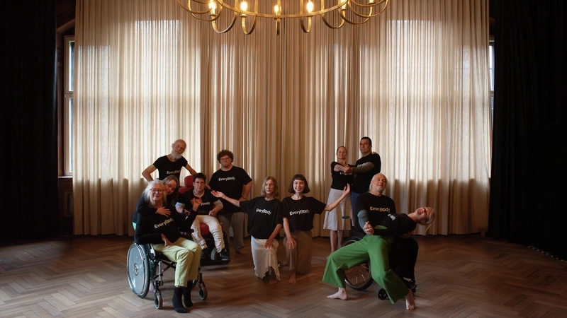 Sieben Frauen und vier Männer haben sich vor einem großen Fenster platziert, das von einem halbtransparenten Vorhang verdeckt wird. Alle tragen schwarze T-Shirts mit der Aufschrift EveryBody. Einige machen Tanzposen, zwei sitzen im Rollstuhl.