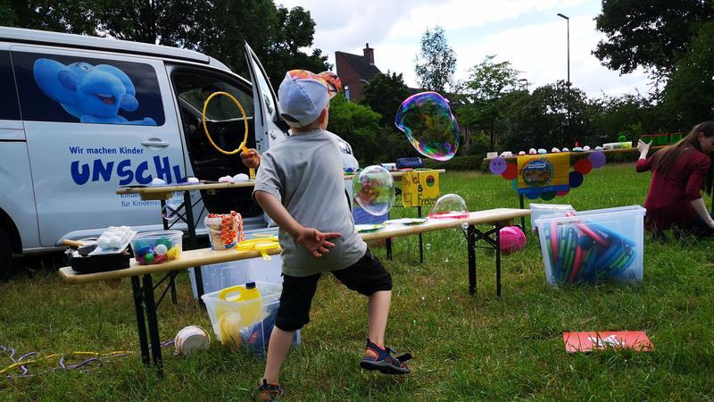 Ein kleiner Junge ist von hinten zu sehen. Er steht auf einer Wiese und schlägt mit einem Federballschläger nach großen Luftblasen. Vor ihm steht eine Bank mit Spielsachen. Dahinter ist ein weißer Transporter zu sehen.