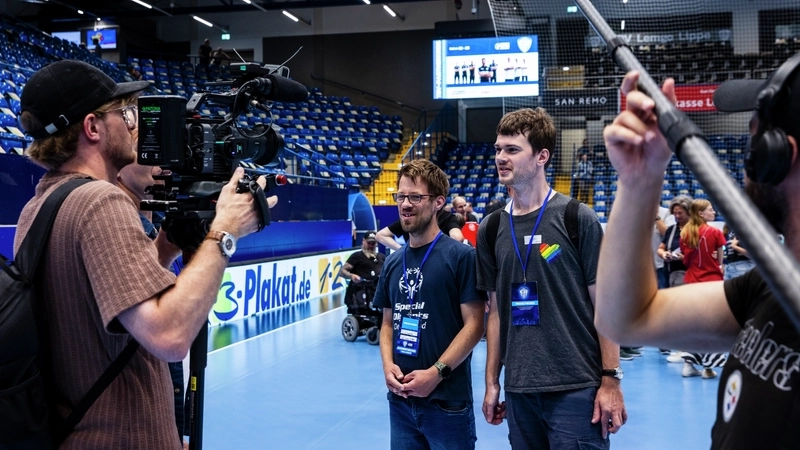 Zwei junge Männer in T-Shirts werden in einer großen Sporthalle mit Zuschauertribühnen interviewt und dabei gefilmt. Sie tragen Identifikationskarten als Anhänger, die sie als Pressevertreter ausweisen.