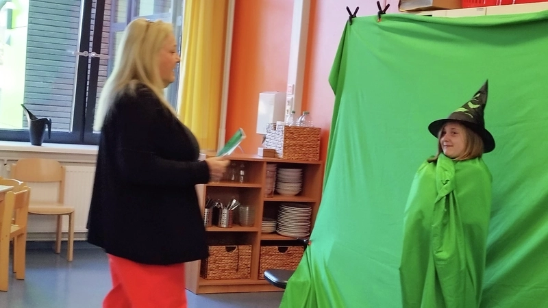 Ein Mädchen steht mit Hexenhut vor  einem grünen Tuch, vor ihr steht eine Frau