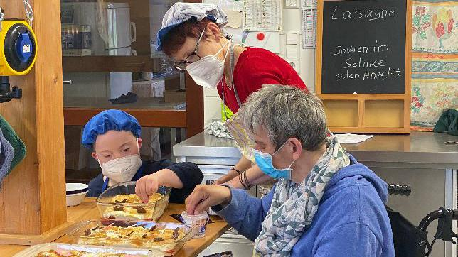 Eine Frau im Rollstuhl, ein Junge mit Downsyndrom und eine Frau arbeiten zusammen in der Küche. Alle tragen Maske.