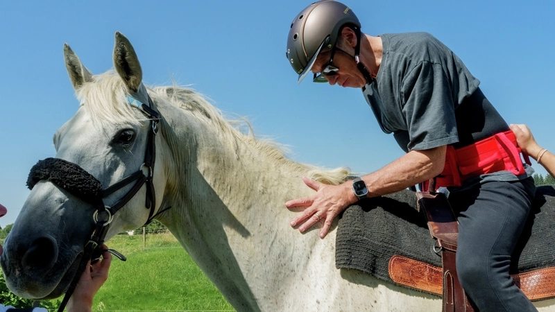Ein älterer Mann mit Helm sitzt auf einem Schimmel. Zwei Personen, von denen man nur die Hände sieht, halten jeweils das Pferd am Zaum und den Mann an einer Schlaufe in seinem Rücken.