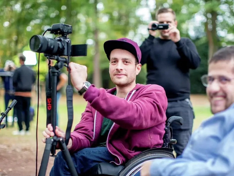 Zwei junge Männer im Rollstuhl bedienen einen Kamera auf einem Stativ.