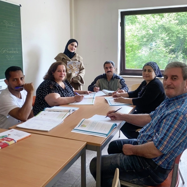 Mehrere Erwachsene siitzen in einem Unterrichtsraum, hinten steht eine junge Frau mit Kopftuch