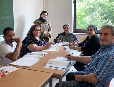 Mehrere Erwachsene siitzen in einem Unterrichtsraum, hinten steht eine junge Frau mit Kopftuch