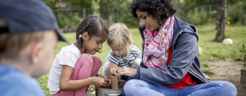 Eine Frau sitzt mit zwei Kleinkindern auf einer Wiese. Sie betrachten etwas in einem Topf, der vor ihnen steht.