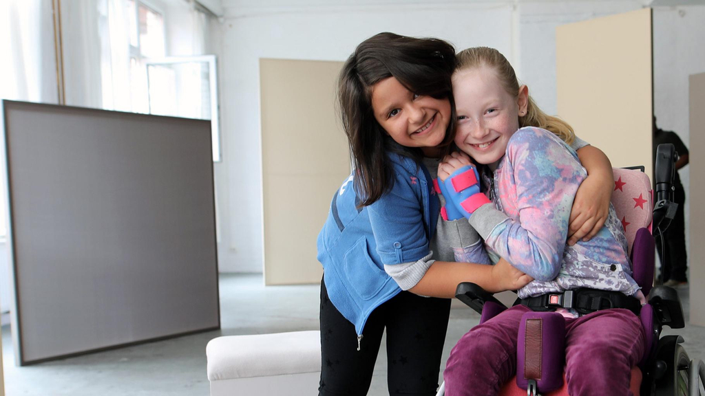 Zwei Mädchen, eines davon im Rollstuhl, umarmen sich und lächeln in die Kamera.