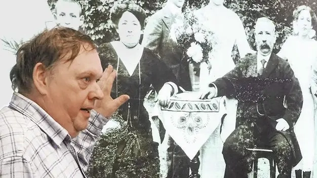 Ein mittelalter Mann in kariertem Hemd steht vor einem s/w-Foto einer Familie und spricht darüber