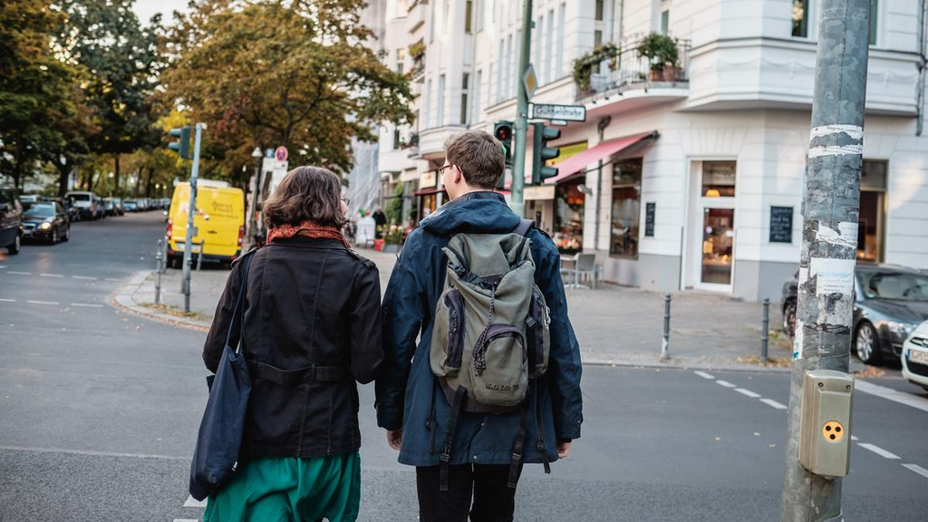 Carlotta und Jasper gehen nebeneinander in der Stadt spazieren. Sie überqueren an einer Ampel eine Straße.