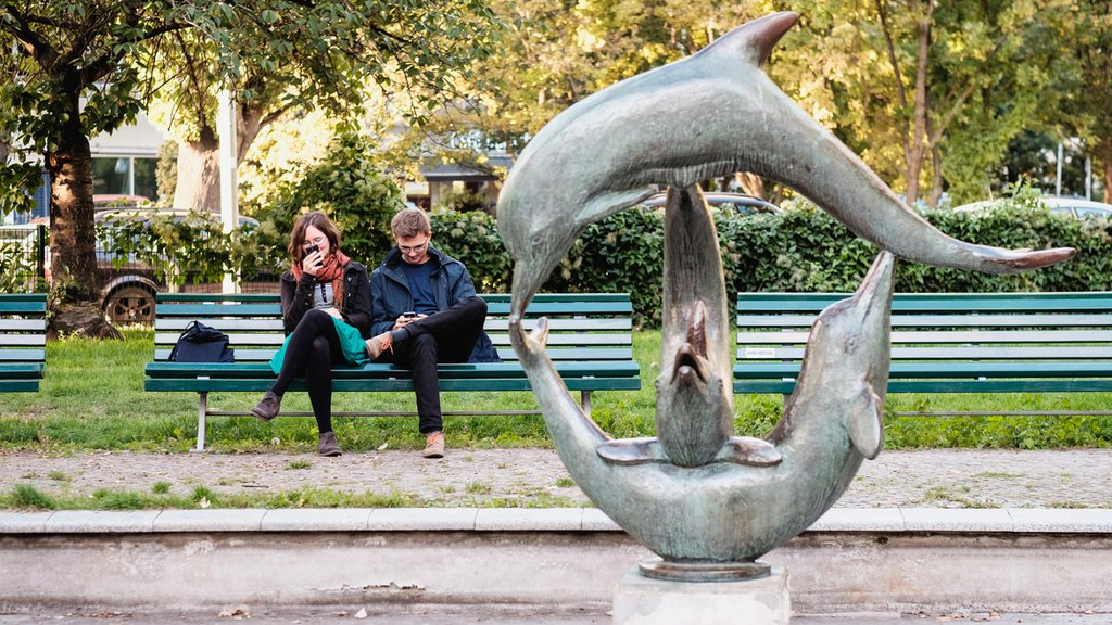 Carlotta und Jasper sitzen im Park auf einer Bank und schauen auf ihre Handys. Im Vordergrund ein Brunnen mit Delfin-Figuren.