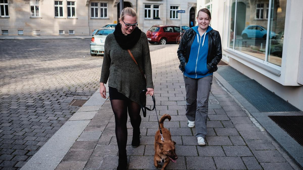 Frauke und Ulrike gehen auf einem Bürgersteig mit dem Hund spazieren.