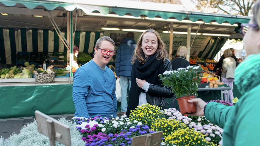 Ein junger Mann und eine junge Frau schauen sich Blumen an einem Marktstand an. Der junge Mann hat das Down-Syndrom.