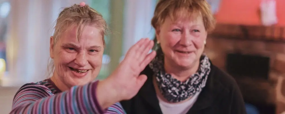 Zwei Frauen mit und ohne Behinderung stehen nebeneinander. Die Frau mit Behinderung hebt lächelnd und abwehrend eine Hand.