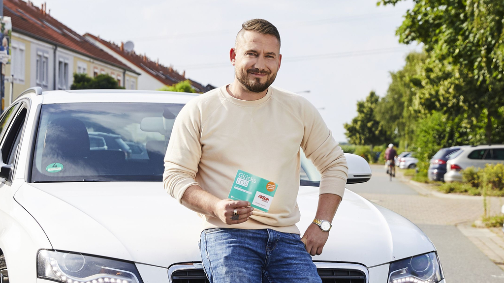 Anton lehnt lässig an einem weißen Auto. Er hält ein Glücks-Los in der Hand.