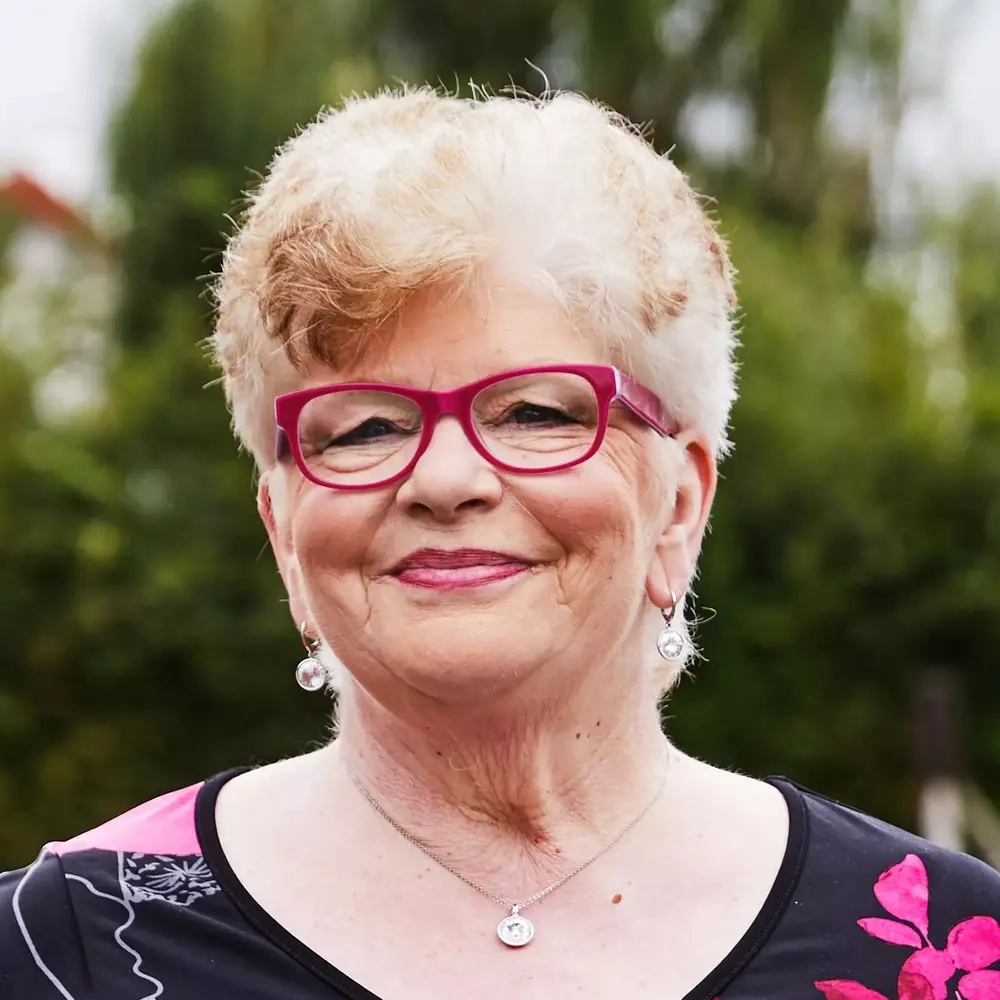 Eine ältere Frau mit kurzem, hellem Haar und pinker Brille lächelt in die Kamera.