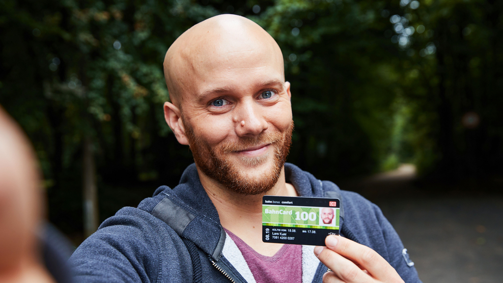 Ein Mann mittleren Alters mit Glatze und Vollbart hält lächeln eine Bahncard in die Kamera.