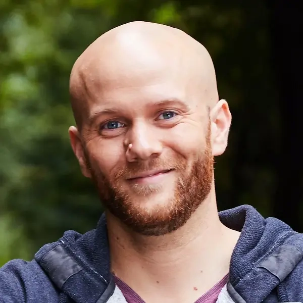 Ein Mann mit Glatze, Vollbart und Kapuzenpulli hält lächelnd ein Glücks-Los in die Kamera.