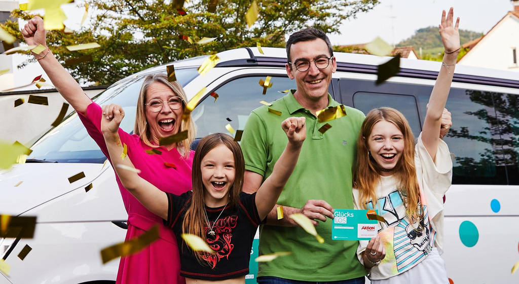 Eine Familie steht im goldenen Konfettiregen. Die Mutter und beiden Töchter recken jubelnd die Arme in die Luft, der Vater hält ein Glückslos in der Hand.
