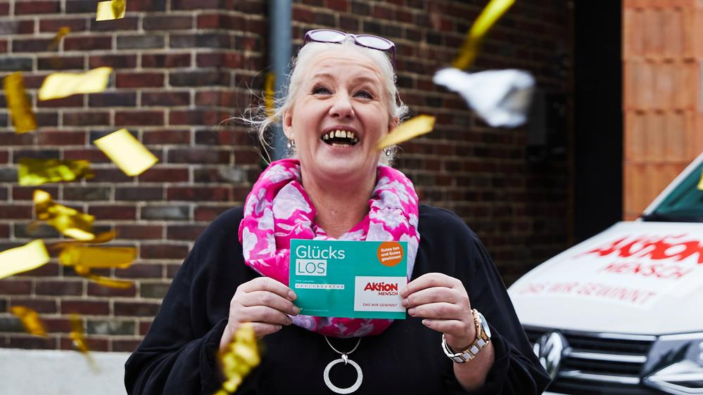 Eine Frau steht lachend in einem goldenen Konfettiregen. Sie hält ein Glücks-Los in der Hand.