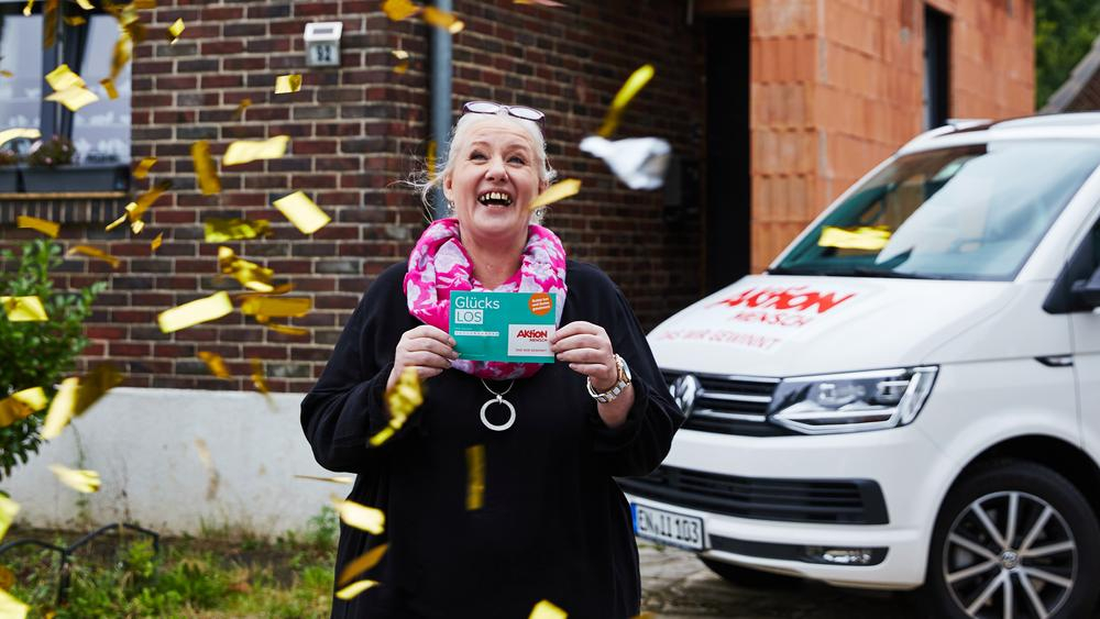 Eine Frau steht lachend in einem goldenen Konfettiregen. Sie hält ein Glücks-Los in der Hand.
