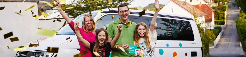 Eine Familie steht im goldenen Konfettiregen. Die Mutter und beiden Töchter recken jubelnd die Arme in die Luft, der Vater hält ein Glückslos in der Hand.