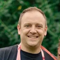 Die Gewinner*innen Bozana und Sebastian stehen Arm in Arm in ihrem Garten. Sie tragen sommerliche Kleidung und halten lächelnd ihr Glückslos in die Kamera. Um ihre Hälse hängen Luftschlangen.