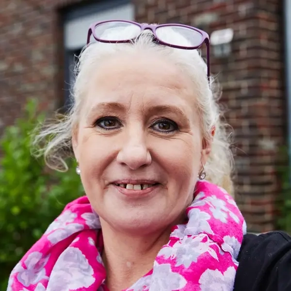 Eine Frau mit blondem Pferdeschwanz und pinkem Halstuch lächelt in die Kamera. Sie steht vor ihrem Haus.