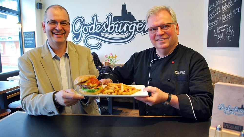 Zwei Männer halten einen Teller mit einem Burger in die Kamera.