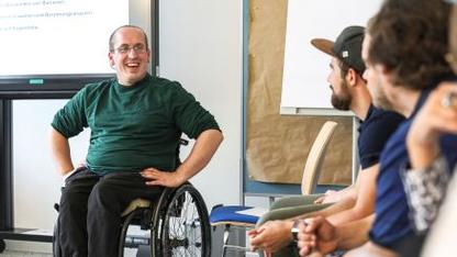 Ein Mann im Rollstuhl sitzt vor einer Gruppe von Menschen und hält einen Vortrag.