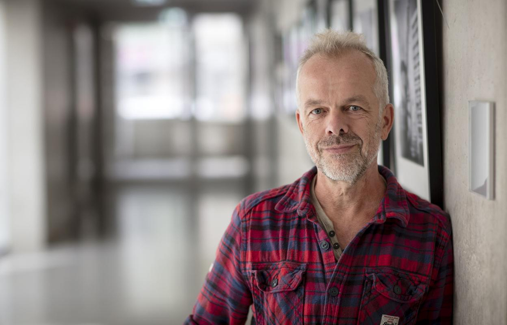 Rainer Tigges steht im Schulflur und lehnt sich an die Wand. Er lächelt in die Kamera.