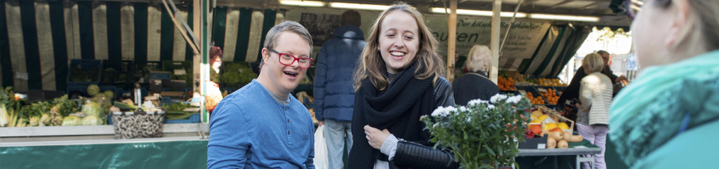 Ein junger Mann und eine junge Frau schauen sich Blumen an einem Marktstand an. Der junge Mann hat das Down-Syndrom.