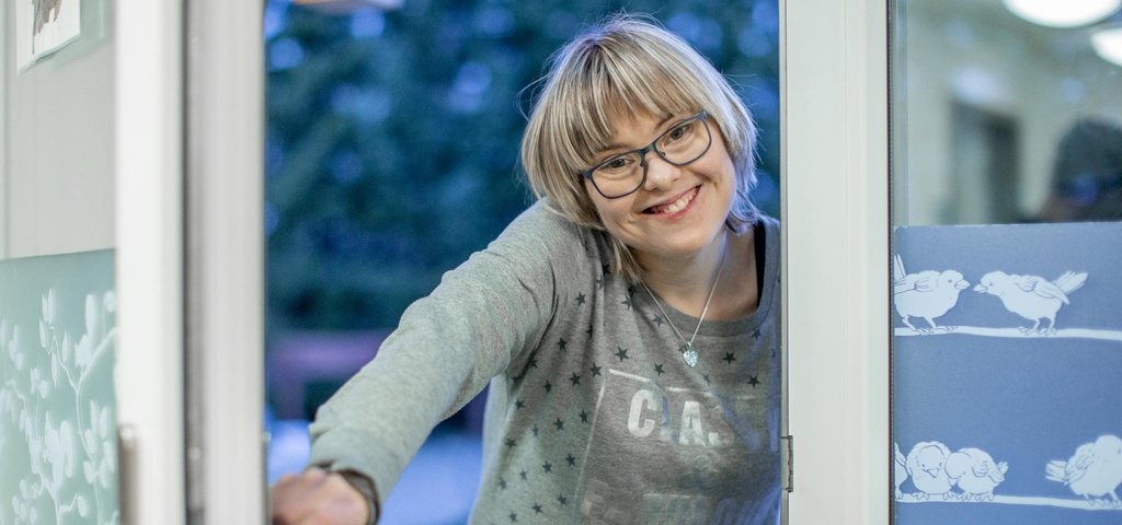 Eine junde Frau öffnet eine Tür und lächelt in die Kamera